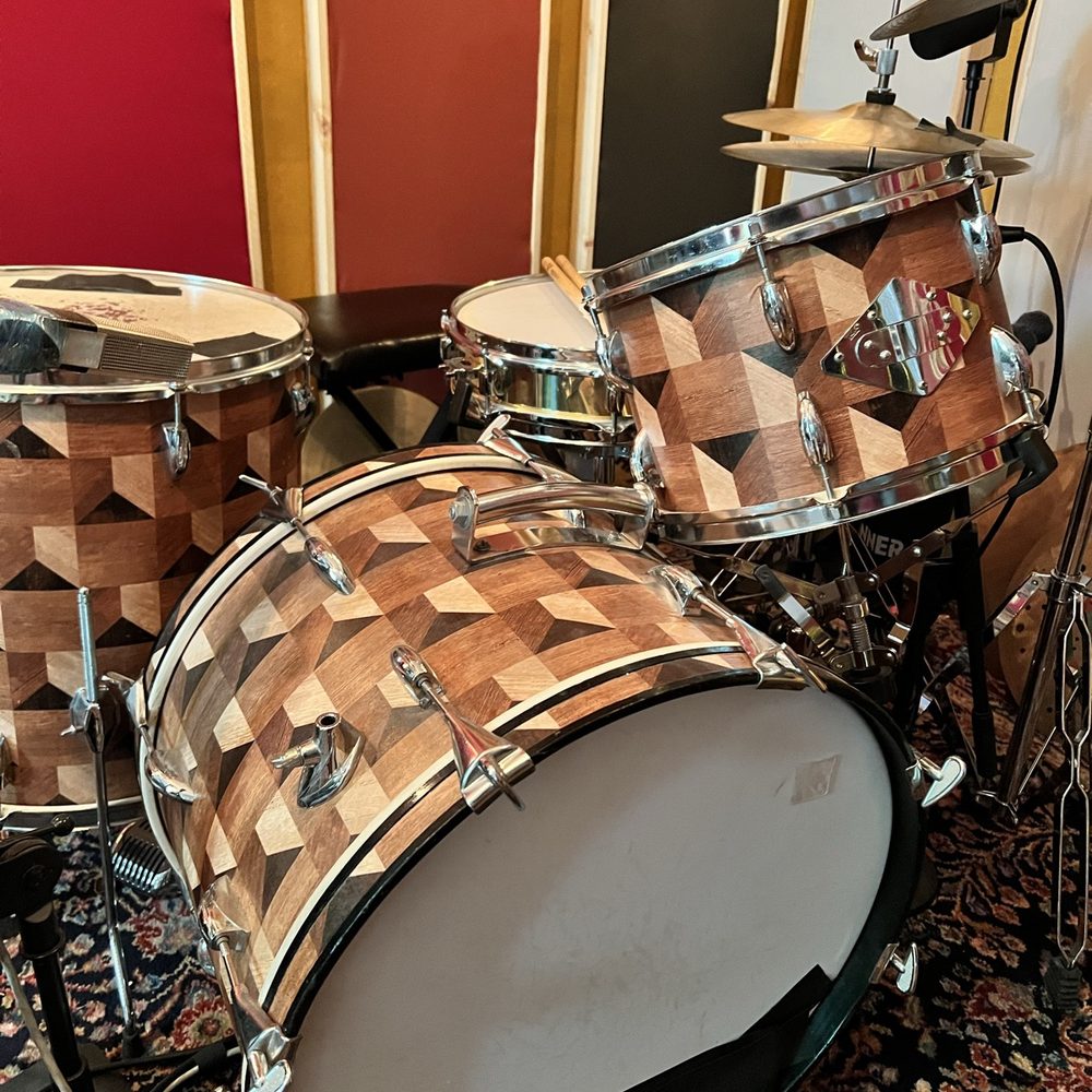 An assembled drum kit shot from above on layered Persian rugs, snare with crossed sticks, a basketweave-finish kick and cymbals
