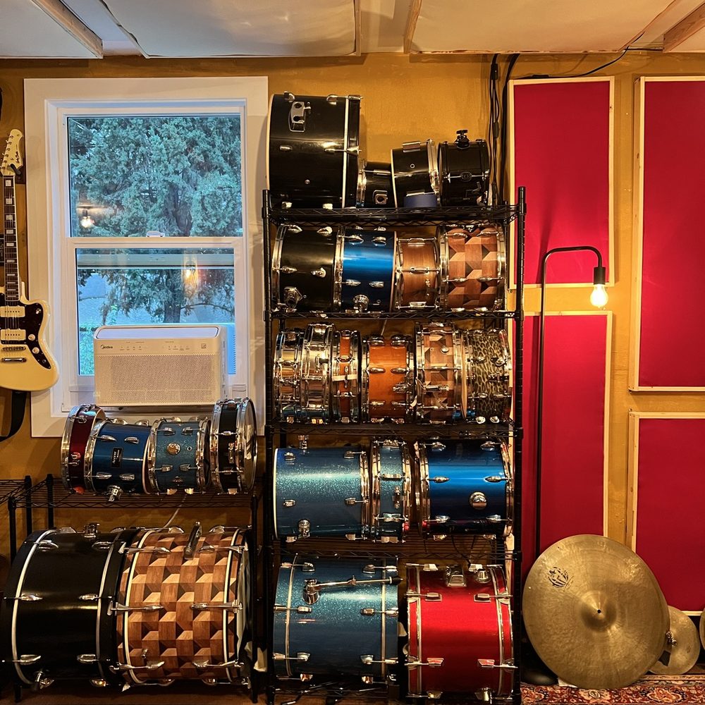 A wall rack stacked with snare and tom drums in blue, red and basketweave finishes beside red acoustic panels and a ride cymbal