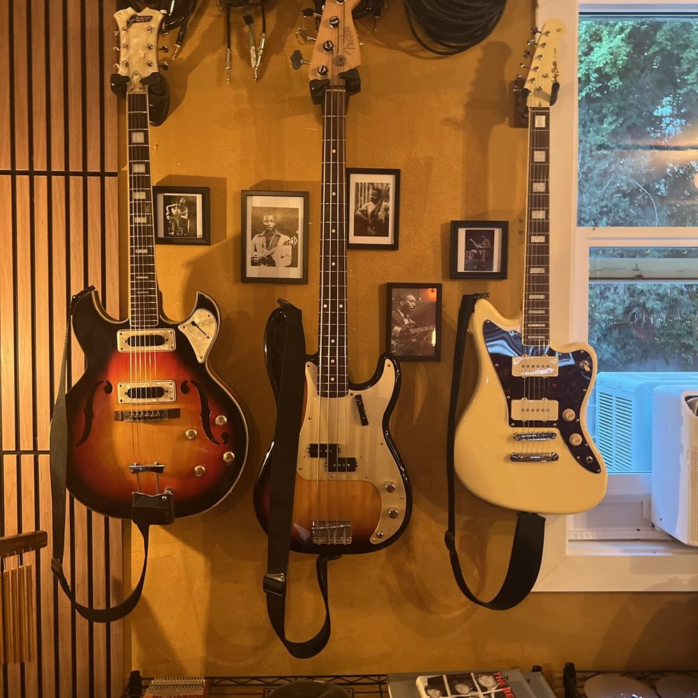 A hollowbody guitar, a sunburst Fender bass and a cream Jazzmaster hung above framed photographs beside a window, coiled cables looped overhead