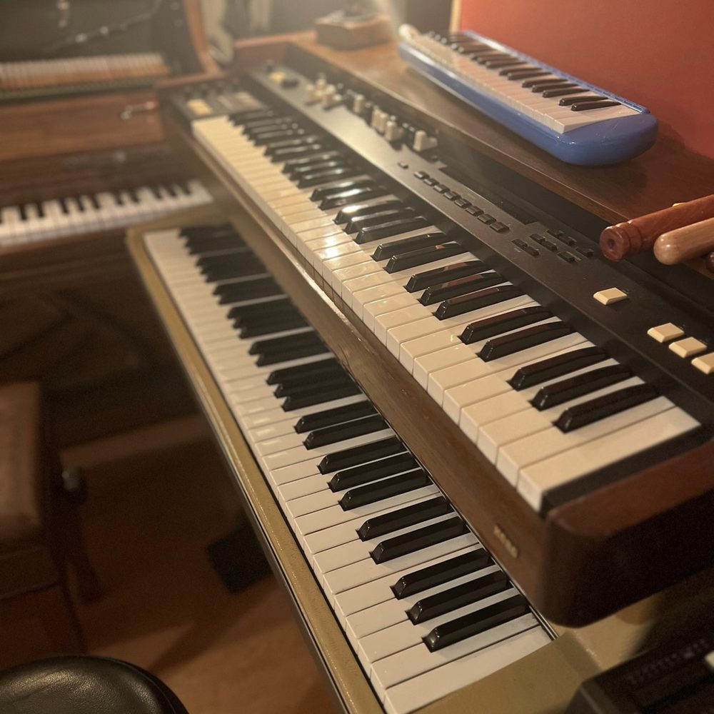 Layered keyboards on a stand: a wooden electric piano, a synthesizer and a blue melodica in warm studio light
