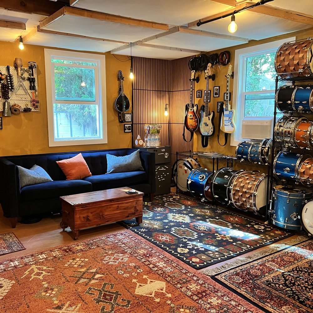 A wide view of the studio lounge: a blue velvet couch and wooden chest on layered rugs facing a wall of guitars and a tall rack of drums