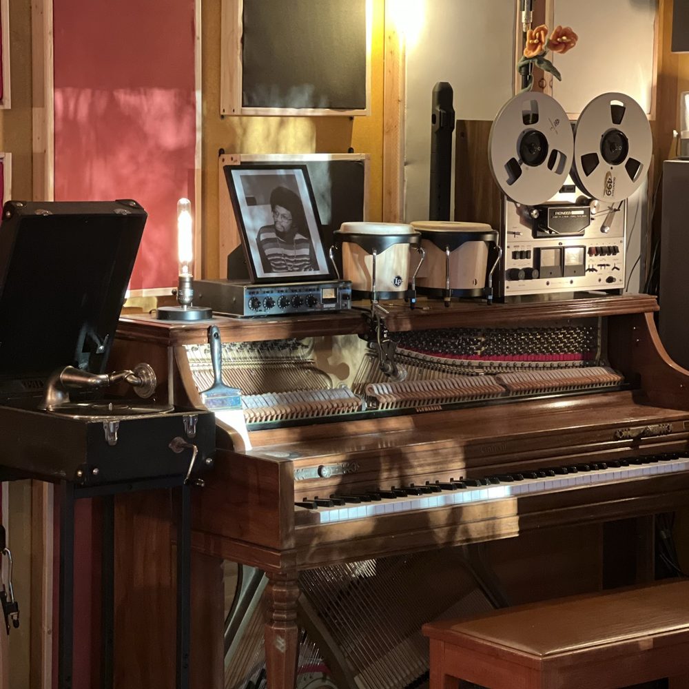 The open action of an upright piano showing its hammers and strings, a wind-up gramophone alongside and a reel-to-reel deck and bongos on top