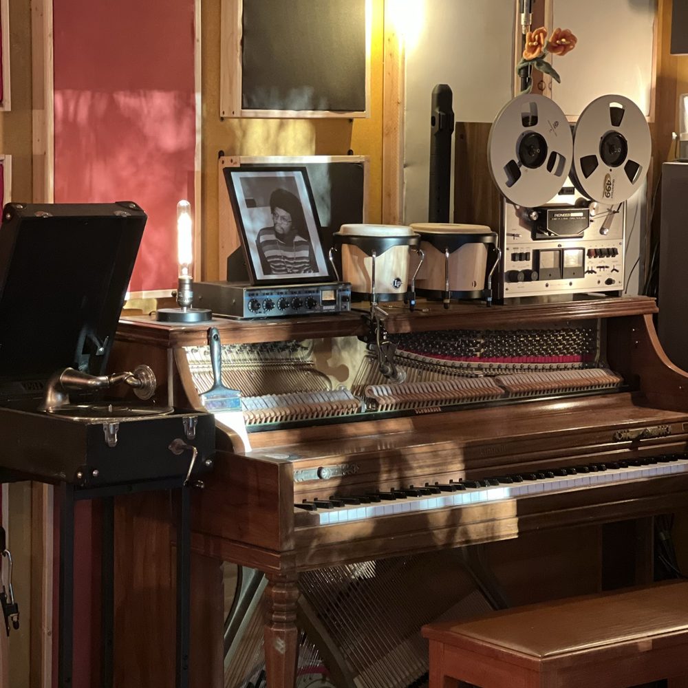 An open upright piano with a silver reel-to-reel tape deck, bongos, a vintage gramophone and a warm Edison bulb on top