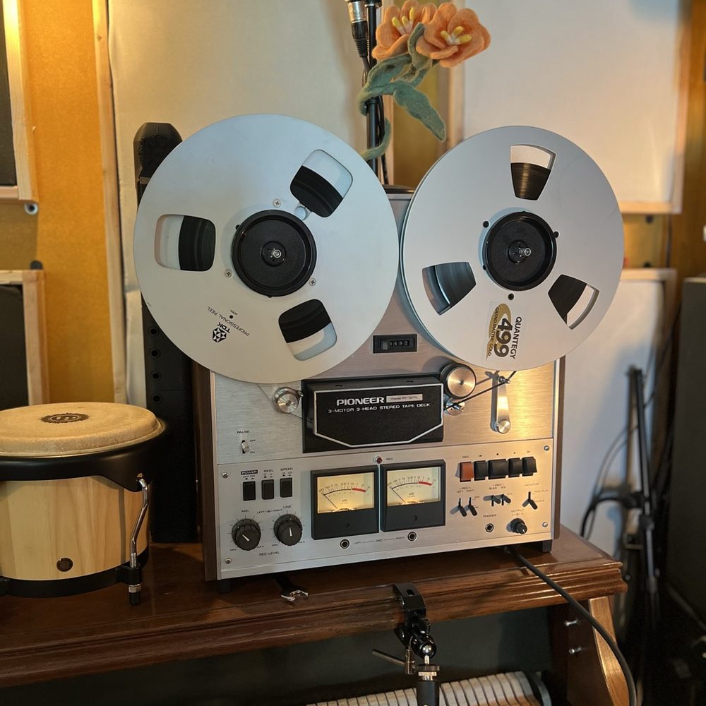 A Pioneer reel-to-reel tape deck with both silver reels loaded, a ribbon microphone above and a wooden bongo alongside